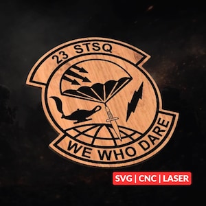 Könnte beinhalten: Holzemblem mit den Texten "23 STSQ" und "WE WHO DARE". Das Design beinhaltet einen Fallschirm, Pfeile, einen Blitz und eine Wunderlampe. Dieses Emblem ist wahrscheinlich für Militär- oder Servicepersonal.