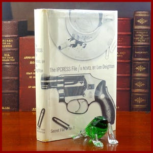May include: A paperback book with a white cover featuring a black and white illustration of a gun and a cigarette in a coffee cup. The book title is "The IPCRESS File" and the author is Len Deighton. The book is part of a series called "Secret File No. 1".