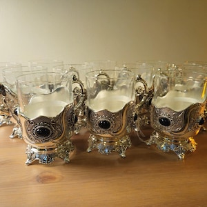 May include: A set of clear glass cups held within ornate silver-toned metal holders. Each holder features intricate scrollwork, a decorative handle, a pedestal base, and a single oval black stone accent embedded on the front of the metal frame.