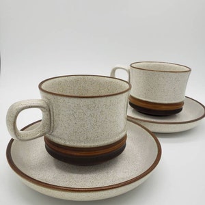 May include: Two white ceramic teacups with brown bands and speckles, each with a matching saucer. The cups and saucers have a speckled glaze.