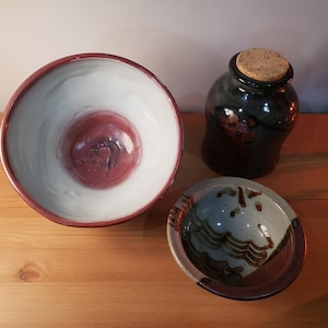 Könnte beinhalten: Drei Keramikstücke: eine große Schüssel mit weißem Innenraum und roter Mitte, eine kleine Schüssel mit braunen und blauen Designs und ein dunkles Gefäß mit Korken. Die Artikel befinden sich auf einer Holzoberfläche.