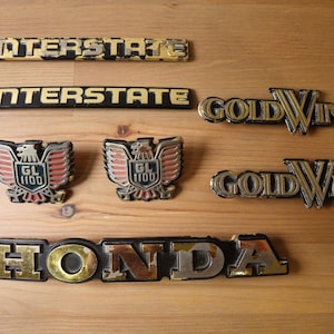 May include: A collection of seven vintage motorcycle emblems including two black and gold INTERSTATE badges, two Gold Wing script emblems, two eagle-shaped GL 1100 badges with red accents, and one large black and gold HONDA nameplate.