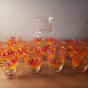 Puede incluir: Una jarra de vidrio transparente y ocho vasos a juego. Cada pieza está decorada con un patrón repetido de diseños de hojas rojas, naranjas y amarillas. El conjunto está dispuesto sobre una superficie de madera.