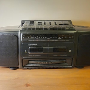 Op de afbeelding: Een zwarte draagbare Magnavox stereocassetterecorder met dubbele cassettedecks gemarkeerd met A en B, een radiotunerdisplay en een opvallende Turbo Bass-luidspreker. Het bovenpaneel bevat diverse bedieningsschakelaars, schuifregelaars en een handvat.