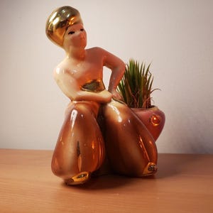 May include: A vintage ceramic planter in the shape of a stylized figure, with a gold-colored head and accents. The figure is holding a small, pink-toned pot with a green plant. The figure's lower body is brown with gold accents.