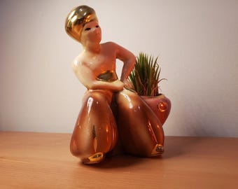 Puede incluir: Una maceta de cerámica vintage con forma de figura estilizada, con cabeza y detalles dorados. La figura sostiene una pequeña maceta de color rosa con una planta verde. La parte inferior del cuerpo es marrón con detalles dorados.