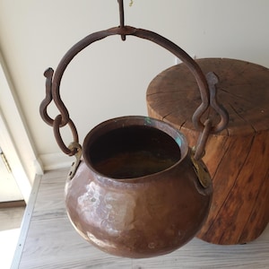 May include: A rustic copper pot with brass accents hangs from a wrought iron hook. The pot is suspended over a wooden stump.