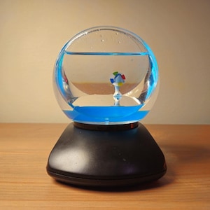 May include: A clear glass globe with blue liquid at the base and a small, colorful decorative element inside. The globe sits atop a black, rounded base. The background is a light beige.