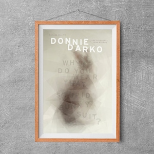 May include: A framed poster with the title "DONNIE DARKO" in large white letters. The poster features text including "Why do you wear that stupid bunny suit?" and the names of the actors. The frame is a light brown colour.