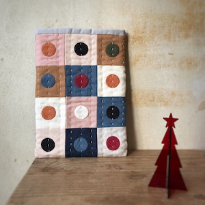 May include: A quilted fabric wall hanging with a geometric pattern of squares. Each square features a different colored circle in the center. The colors include brown, pink, white, blue, red, and green.