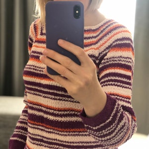 Pu&ograve; includere: Un maglione in maglia a maniche lunghe con righe orizzontali nei toni del viola scuro, arancione brillante e bianco sporco. Il capo presenta uno scollo rotondo e un motivo a maglia testurizzato su tutto il corpo e le maniche.