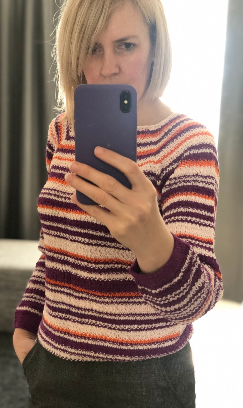 Pu&ograve; includere: Un maglione in maglia a maniche lunghe con righe orizzontali nei toni del viola scuro, arancione brillante e bianco sporco. Il capo presenta uno scollo rotondo e un motivo a maglia testurizzato su tutto il corpo e le maniche.