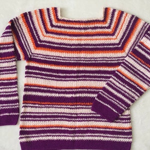 Pu&ograve; includere: Un maglione in maglia a maniche lunghe con scollo quadrato e un motivo a righe orizzontali nei toni del viola intenso, arancione brillante e bianco sporco. Il capo presenta una consistenza lavorata a maglia uniforme su tutto il corpo e le maniche.