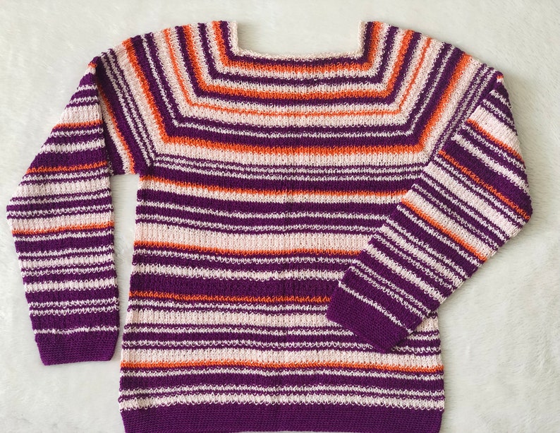 Pu&ograve; includere: Un maglione in maglia a maniche lunghe con scollo quadrato e un motivo a righe orizzontali nei toni del viola intenso, arancione brillante e bianco sporco. Il capo presenta una consistenza lavorata a maglia uniforme su tutto il corpo e le maniche.