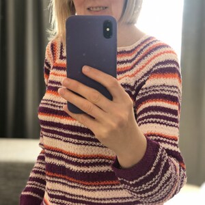 Pu&ograve; includere: Un maglione in maglia a maniche lunghe con righe orizzontali nei toni del viola scuro, arancione brillante e bianco sporco. Il capo presenta uno scollo rotondo e polsini a coste, con un motivo a maglia strutturato e uniforme su tutto il tessuto.
