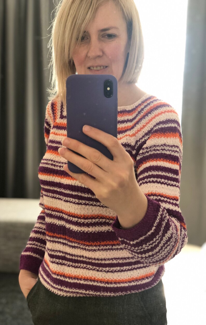 Pu&ograve; includere: Un maglione in maglia a maniche lunghe con righe orizzontali nei toni del viola scuro, arancione brillante e bianco sporco. Il capo presenta uno scollo rotondo e polsini a coste, con un motivo a maglia strutturato e uniforme su tutto il tessuto.
