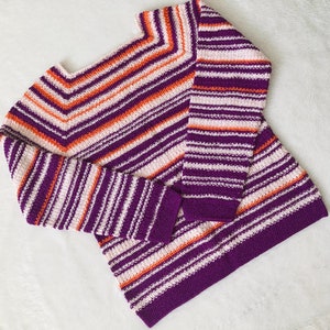 Pu&ograve; includere: Un maglione a maniche lunghe caratterizzato da un motivo a righe orizzontali nei toni del viola intenso, arancione brillante e bianco sporco. Il capo presenta uno scollo quadrato e una trama lavorata a maglia, con righe a motivo chevron.