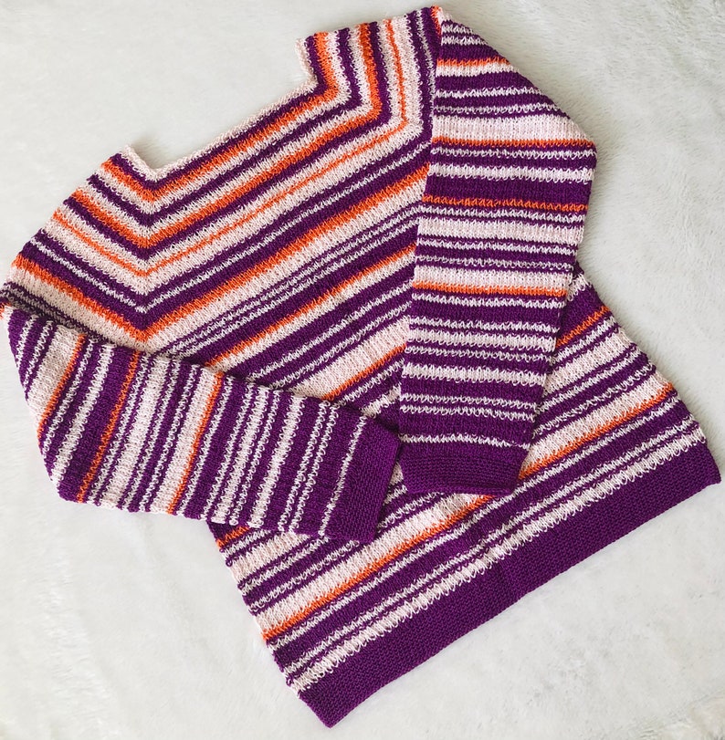 Pu&ograve; includere: Un maglione a maniche lunghe caratterizzato da un motivo a righe orizzontali nei toni del viola intenso, arancione brillante e bianco sporco. Il capo presenta uno scollo quadrato e una trama lavorata a maglia, con righe a motivo chevron.