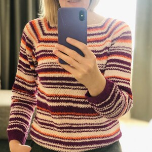 Pu&ograve; includere: Un maglione in maglia a maniche lunghe con righe orizzontali nei toni del crema, viola scuro e arancione brillante. Il capo presenta uno scollo rotondo e polsini a coste, con un motivo a righe costante che si estende su maniche e busto.