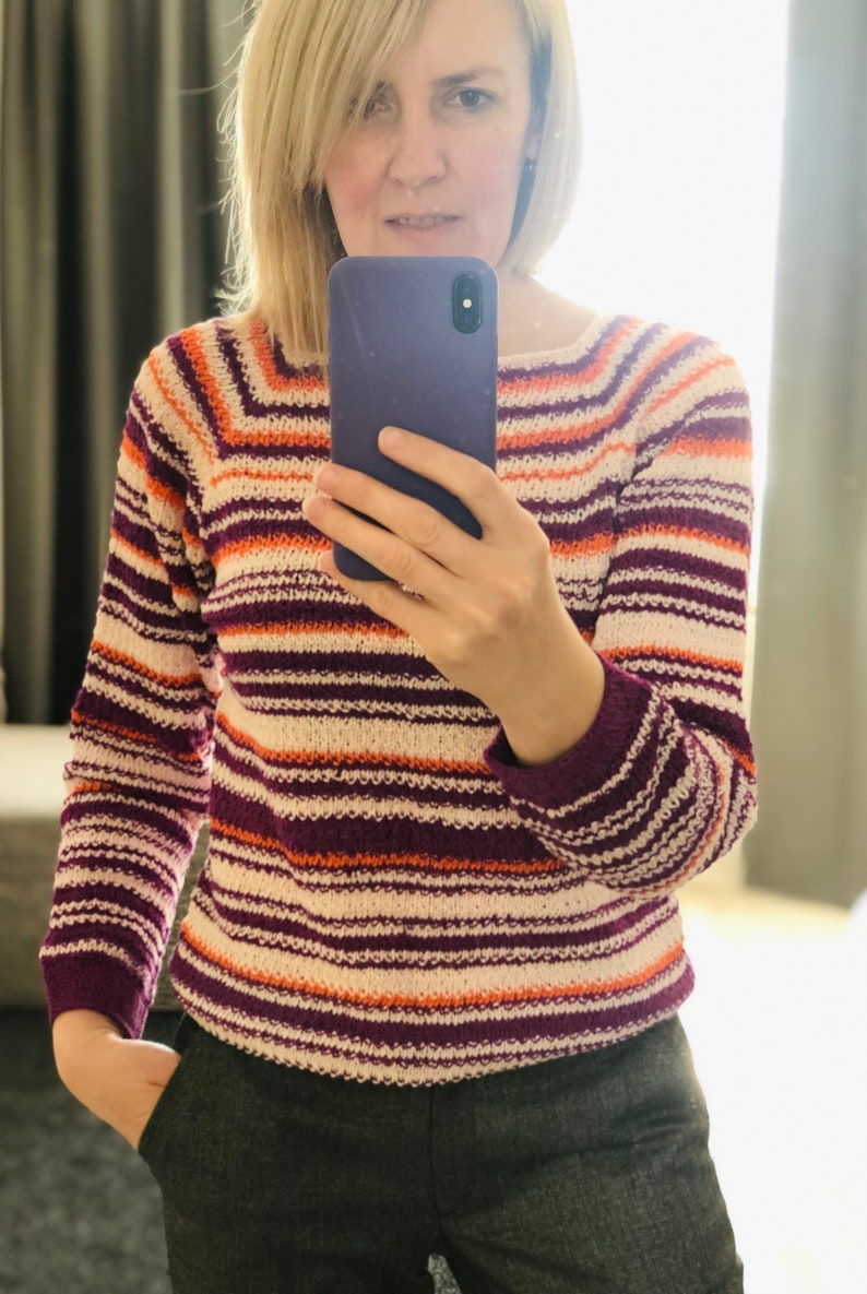 Pu&ograve; includere: Un maglione in maglia a maniche lunghe con righe orizzontali nei toni del crema, viola scuro e arancione brillante. Il capo presenta uno scollo rotondo e polsini a coste, con un motivo a righe costante che si estende su maniche e busto.