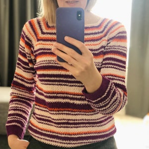 Pu&ograve; includere: Un maglione in maglia a maniche lunghe con righe orizzontali nei toni del viola scuro, arancione brillante e bianco sporco. Il capo presenta uno scollo a barchetta e polsini a coste, con un motivo a righe costante su maniche e busto.