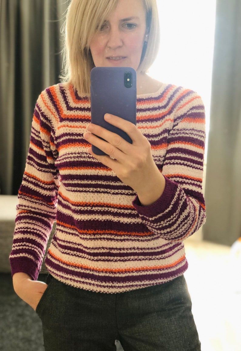 Pu&ograve; includere: Un maglione in maglia a maniche lunghe con righe orizzontali nei toni del viola scuro, arancione brillante e bianco sporco. Il capo presenta uno scollo a barchetta e polsini a coste, con un motivo a righe costante su maniche e busto.