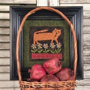 May include: A woven wicker basket filled with red pomegranates. The basket has a brown and tan woven pattern. The basket is sitting in front of a framed picture of a cat with flowers.