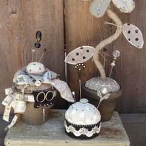 May include: Three fabric pincushions in the shape of flowers. The largest pincushion is a white and brown flower with a brown button center. The other two pincushions are smaller and have a white fabric base with a brown trim.