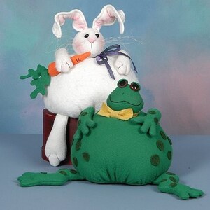May include: A white plush bunny holding a carrot sits on a wooden stand next to a green plush frog wearing a yellow bow tie.