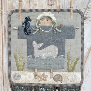 May include: A fabric wall hanging with a sea theme. It features a whale, seahorse, and seashells. The text "By the Sea" is stitched at the bottom. The piece is held by wooden clothespins and has a gray and blue color scheme.