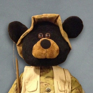 May include: A plush black bear wearing a brown and tan fishing vest and a brown hat with a fishing rod. The bear has large brown eyes and a brown nose.