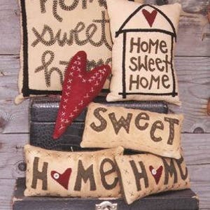 May include: A stack of five decorative pillows with a home sweet home theme. The pillows are made of beige fabric with brown stitching and feature the words "Home Sweet Home" in various styles. The pillows are arranged on a black suitcase with a silver latch.