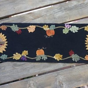 May include: Black oval table runner with a fall theme. Features sunflowers, pumpkins, and autumn leaves in shades of orange, yellow, and green. The runner is approximately 36 inches long.