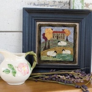 May include: A framed needlepoint artwork depicting a farmhouse with a green roof and yellow walls, surrounded by trees with orange and yellow leaves.  Four white sheep are grazing in a field in front of the house.