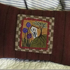 May include: A maroon pillow with a brown and beige checkered border surrounding a square with a red, yellow, and green embroidered design of a small house with a tree and purple flowers.