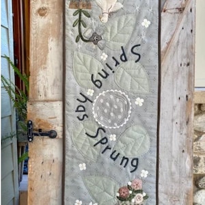 May include: A gray and green fabric wall hanging with the text "Spring has Sprung" and a white rabbit pulling a wagon with flowers. The background is a light blue color with white flowers.