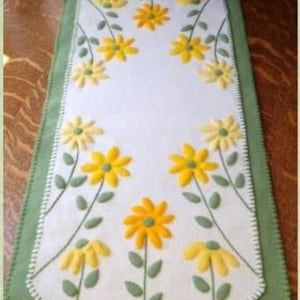 May include: A white felt table runner with a green border and yellow flower embroidery. The runner features a repeating pattern of yellow flowers with green stems and leaves. The text "2018 Cath's Pennies Designs" is embroidered in the bottom center of the runner.