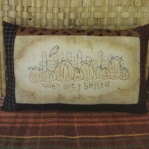May include: A beige pillow with a black and white plaid border. The pillow features an embroidered design of pumpkins, a fence, a bird, and the text "We are blessed".