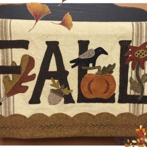 May include: A beige fabric wall hanging with the word "FALL" in black felt letters. The letters are decorated with felt appliques of leaves, acorns, a pumpkin, a crow, and a flower.