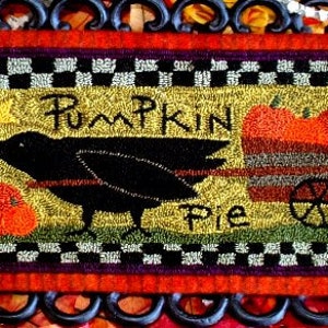 May include: A black crow with a yellow star above it pulls a wagon with a pumpkin pie. The wagon has a wheel and is on a yellow background with a black and white checkered border. The text "Pumpkin Pie" is stitched in brown thread.