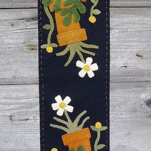 May include: A black felt wall hanging with a stitched design of stacked potted plants with green leaves and white flowers. The pots are brown and have the letters "XXX" stitched on them.