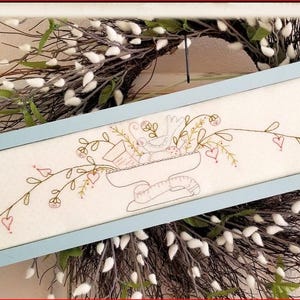 May include: A rectangular, light blue framed artwork featuring an embroidered design. The artwork displays a central floral arrangement with a bird, and a vine with hearts and leaves. The background is a cream color. The artwork is set against a white and brown wreath.