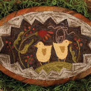 May include: Oval-shaped, hand-stitched rug featuring two yellow chicks, flowers, and a bug on a dark brown background. The rug has a starburst border and a brown and tan plaid outer edge. The rug is displayed in a woven basket with green moss.