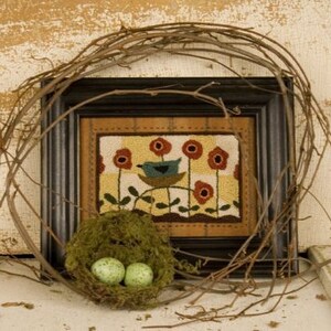 May include: A framed needlepoint artwork featuring a blue bird perched on a branch with red flowers and a yellow background. A bird's nest with two green eggs sits in front of the frame.