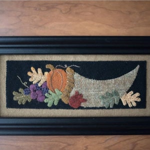 May include: A framed needlepoint artwork depicting a cornucopia overflowing with autumnal elements, including a pumpkin, grapes, and leaves. The cornucopia is stitched in beige and brown wool, while the pumpkin is orange, the grapes are purple, and the leaves are green, brown, and red.
