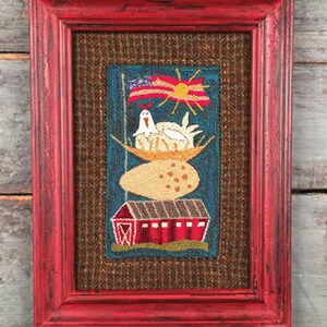 May include: A framed needlepoint artwork depicting a red barn, a large egg, a hen sitting on a nest of eggs, and a sun with a small American flag. The artwork is framed in a red wood frame.