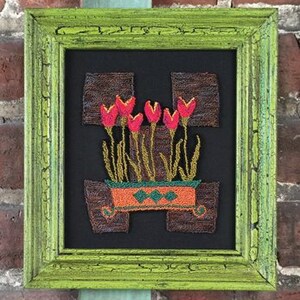 May include: A framed artwork featuring a colorful embroidered design of red flowers in a pot with a geometric pattern. The frame is green with a distressed finish.