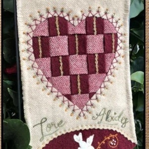 May include: A beige fabric wall hanging with a red and brown heart in the center. The heart is stitched with a brown thread and has a checkered pattern. The words "Love Abides" are stitched in green thread below the heart. A red fabric with a white dove and a brown vine is stitched at the bottom of the hanging.