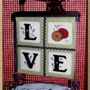 May include: A framed felt wall hanging with the word "LOVE" spelled out in black letters. Each letter is surrounded by a green border and has small embroidered flowers in red, blue, and white. The frame is black with a decorative scroll design. The hanging is displayed on a black metal stand with a white crocheted doily on the bottom shelf.