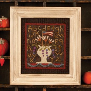 May include: A framed needlepoint piece with a white vase filled with flowers and a small American flag. The background is brown with the letters of the alphabet embroidered around the vase.